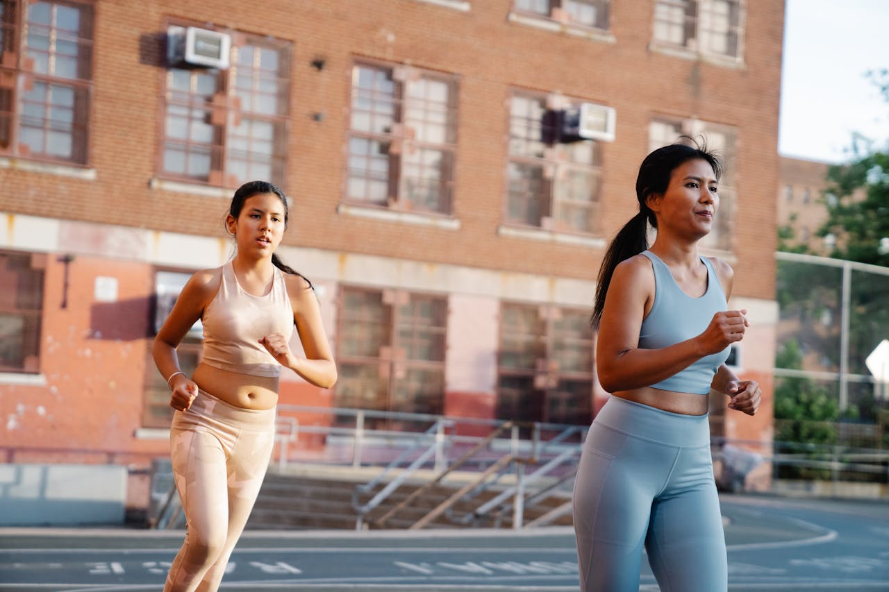 Two women jogging on an outdoor court near a brick building in activewear.